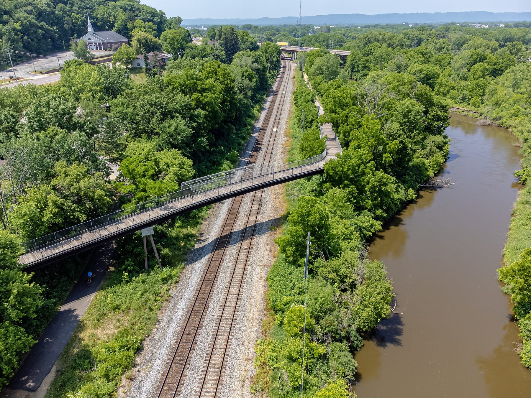 An aerial view shows the La Crosse River curving through a green corridor beside rail tracks and a trail bridge.