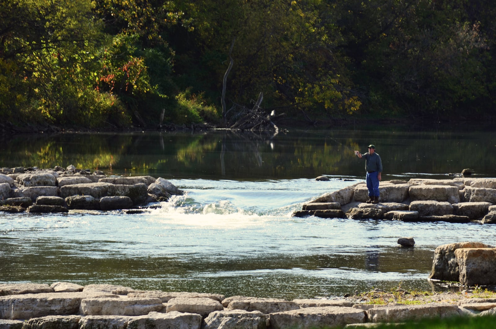 A person fishes from a rock in the Maquoketa River, with wooded banks and a shallow rocky channel around them.