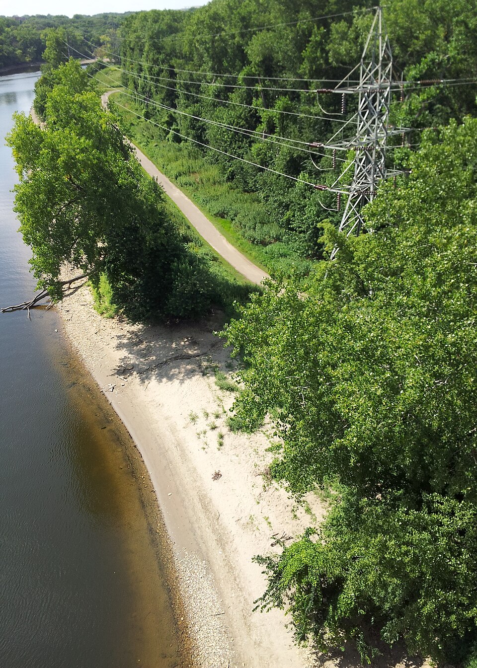 An aerial view of the Mississippi River shoreline at Hidden Falls in Saint Paul, with a sandy bank, riverside trail, trees, and power lines.
