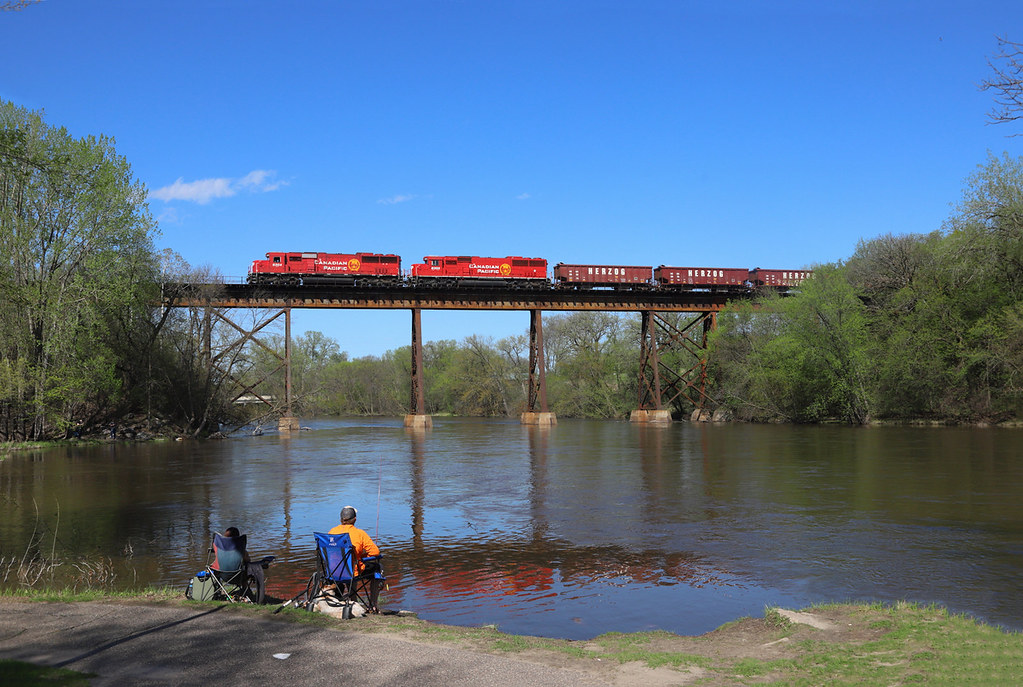 The Crow River flows under a railroad bridge in Rockford, Minnesota, with people sitting near the water at the landing.
