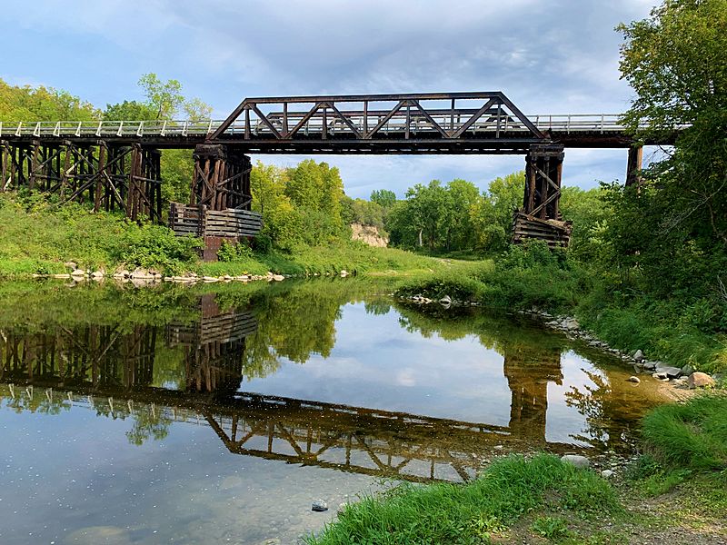 The Red Lake River reflects a railroad bridge, green banks, and cloudy sky in calm water.