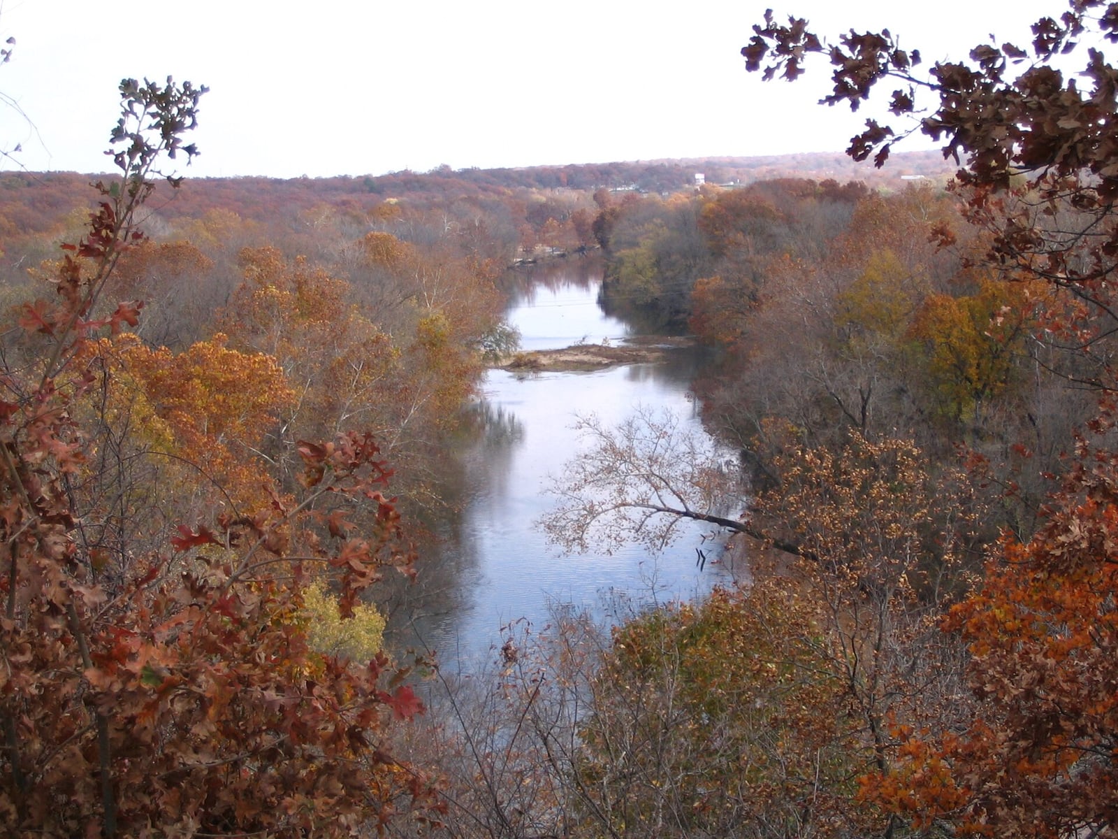 Shoal Creek bends below a wooded overlook near Joplin with a shallow riffled channel visible through the trees.