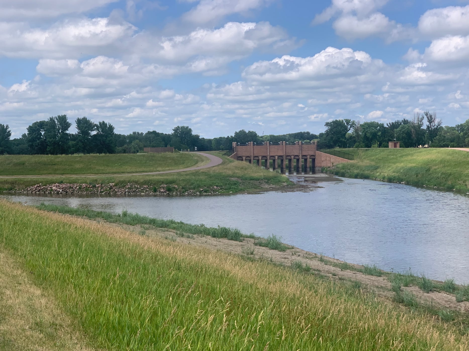Skunk Creek meets the Big Sioux River in Sioux Falls below grassy banks and a cloudy sky.