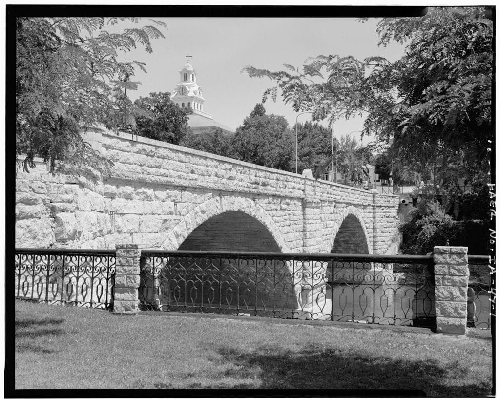 Elkader Keystone Bridge crosses the Turkey River with stone arches, town buildings, and riverbank trees in view.